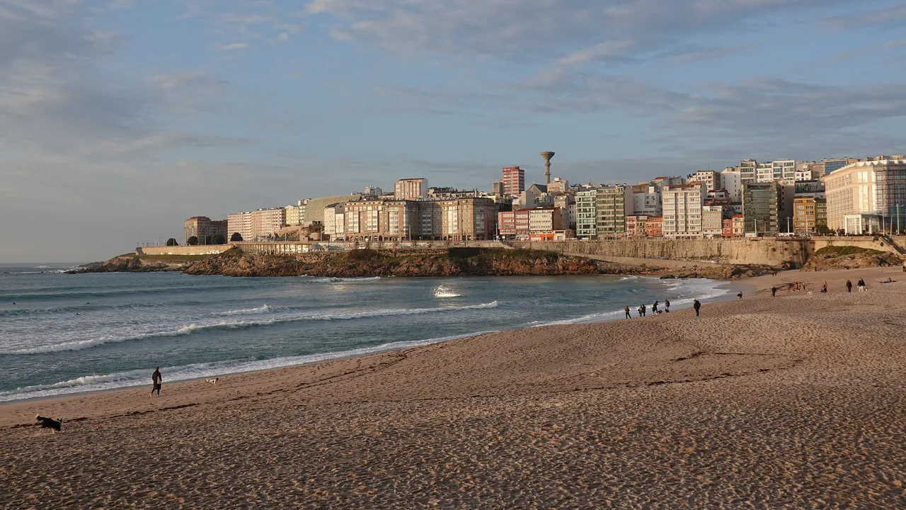La playa del Orzán al atardecer, con las olas rompiendo, gente andando por la arena y la ciudad entera levantada justo detrás.