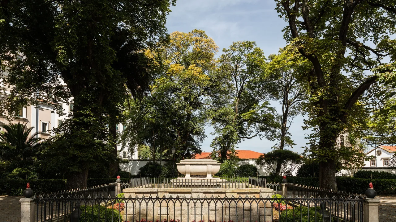 El interior del Jardín de San Carlos, cerrado por olmos altos, con la balaustrada de piedra y los tejados rojos de la Ciudad Vieja asomando detrás.