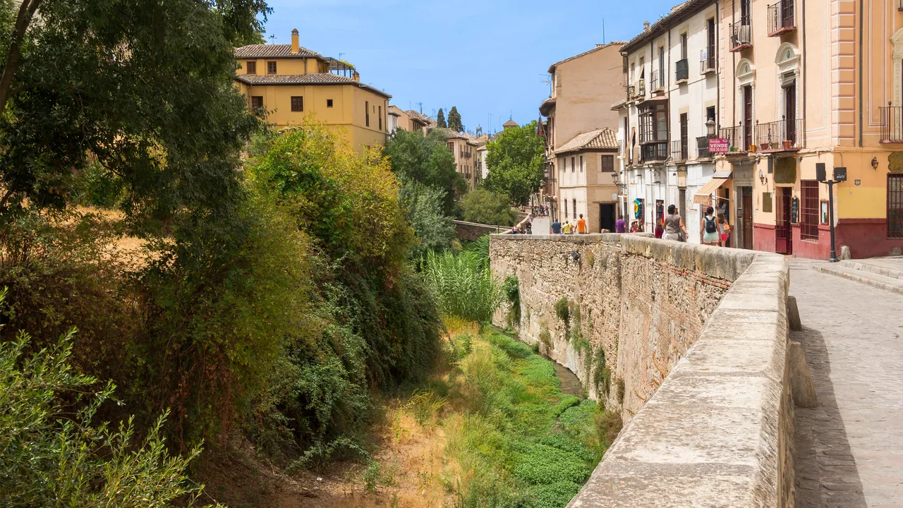 La Carrera del Darro: el paseo empedrado llano junto al pretil de piedra, el cauce del río con vegetación a un lado y las casas antiguas subiendo al otro.