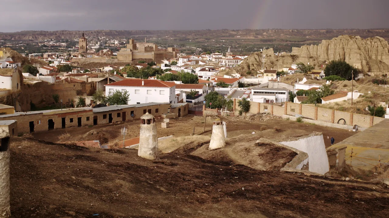 El barrio de las Cuevas de Guadix desde arriba: chimeneas blancas encaladas en primer plano, las casas del casco y la torre de la catedral al fondo, y los cerros de arcilla a la derecha.