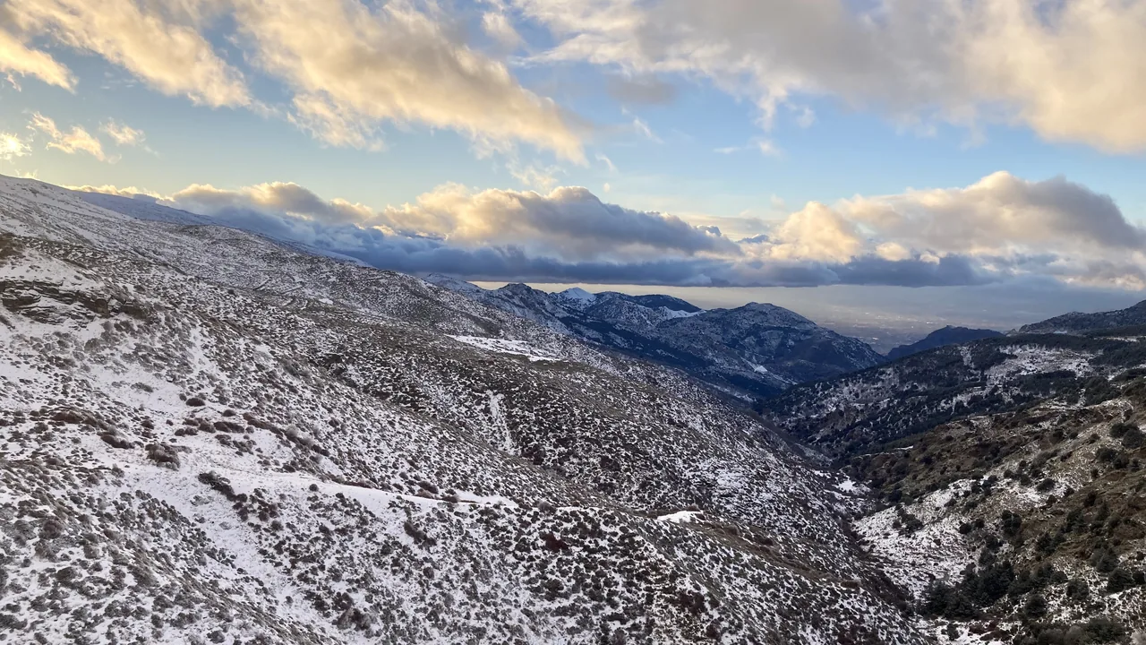 La ladera de Sierra Nevada cubierta de nieve al atardecer, con el valle bajando hacia la vega y una banda de nubes iluminadas sobre las cumbres.