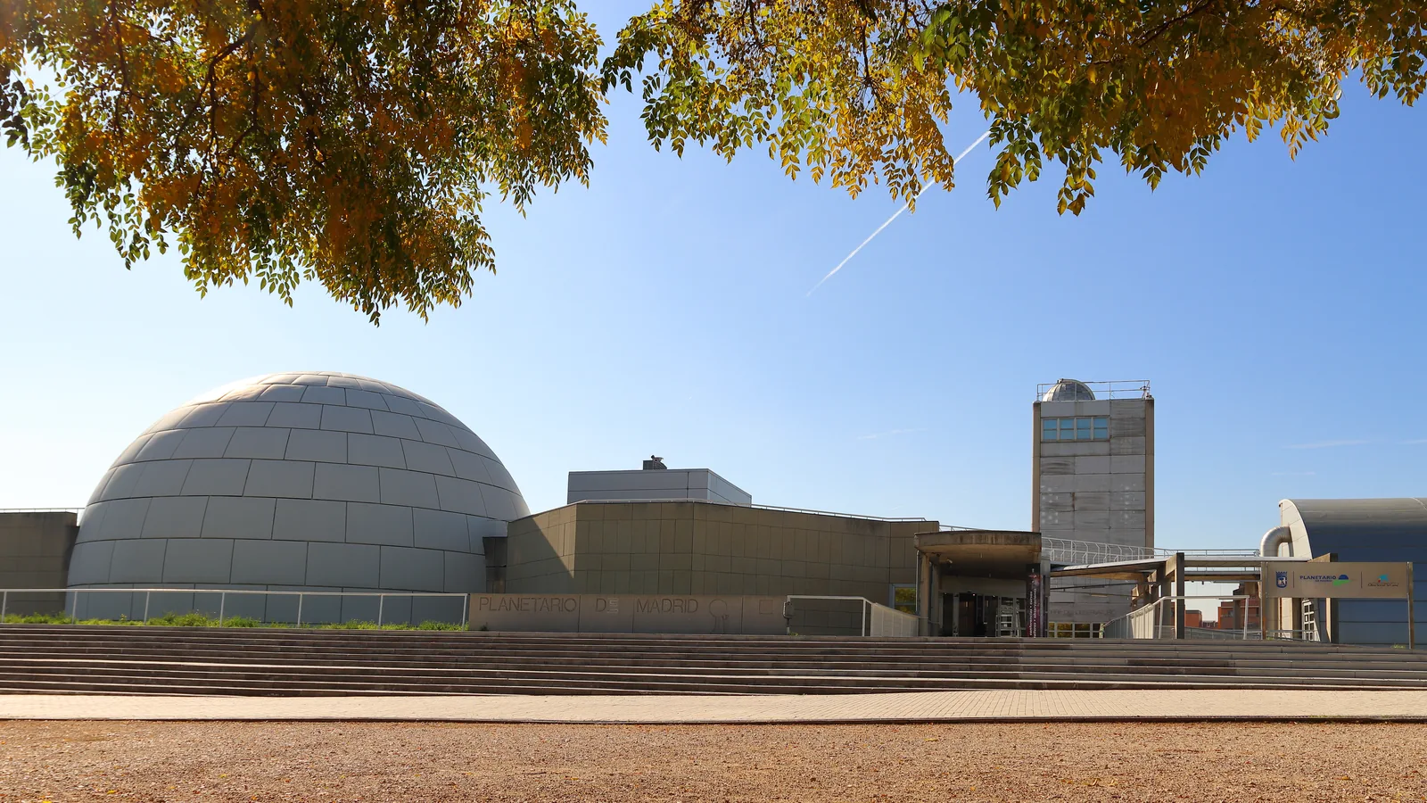 Cúpula gris del Planetario de Madrid con su rótulo grabado, enmarcada por ramas de otoño en el parque Tierno Galván.