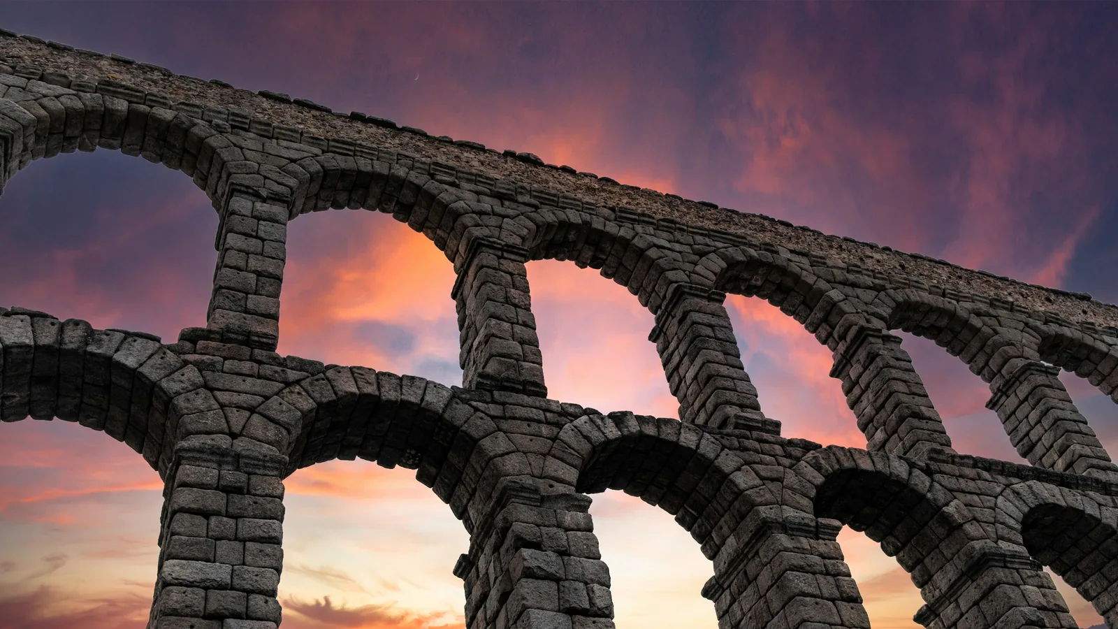 El acueducto de Segovia en contrapicado al atardecer, con el cielo naranja asomando entre los arcos.