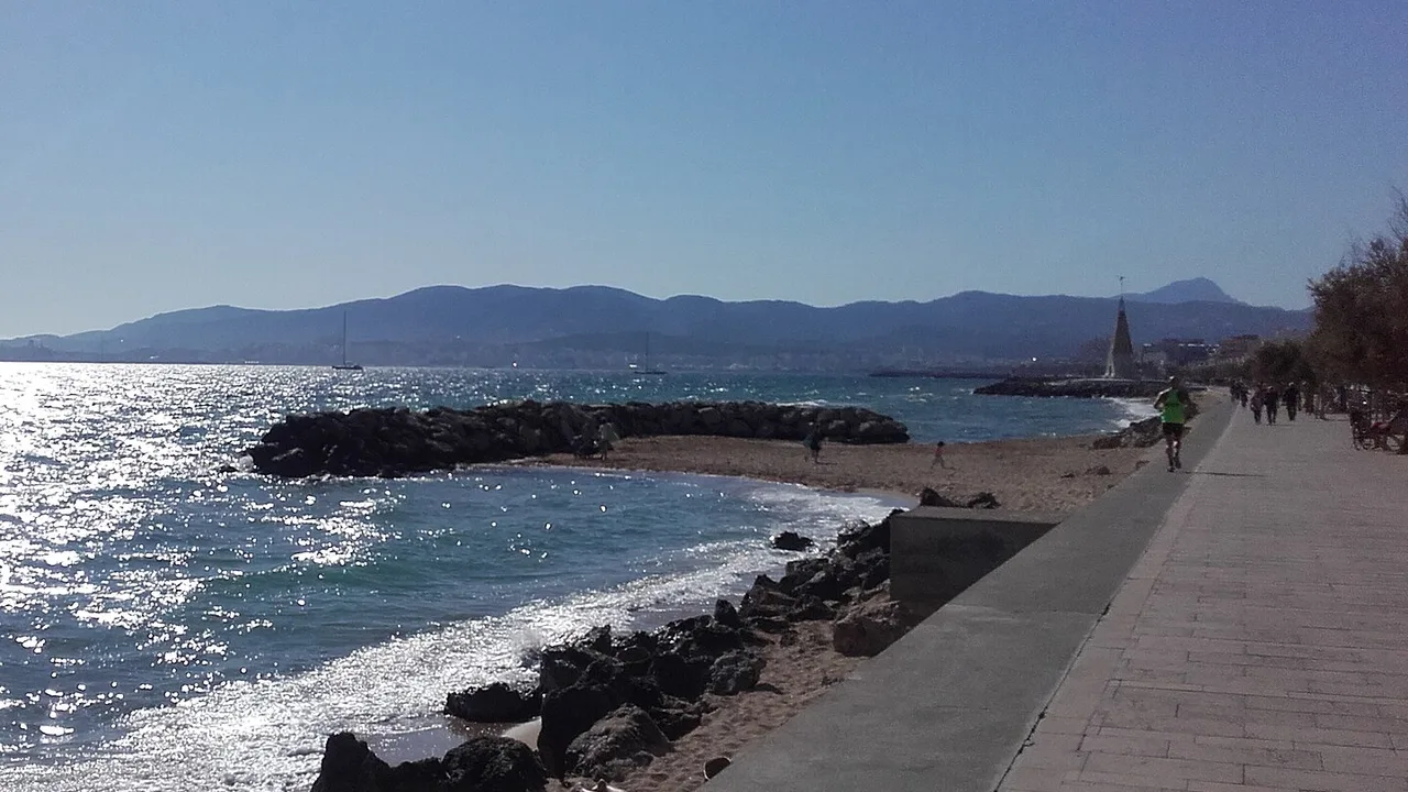 El paseo marítimo del este de Palma, llano y pegado al agua, con la cala, el espigón y la sierra al fondo.