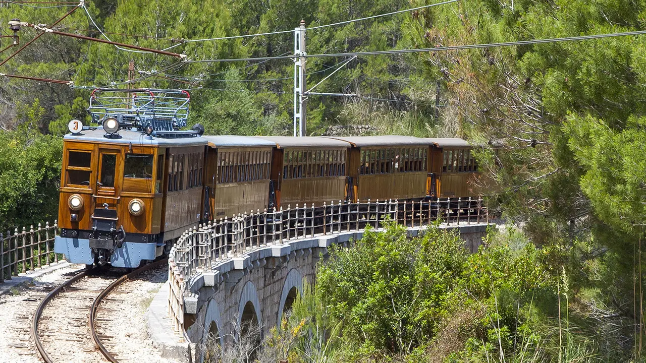 El tren de madera del Ferrocarril de Sóller cruzando un viaducto de piedra, con la ladera de pinos de la Tramuntana detrás.