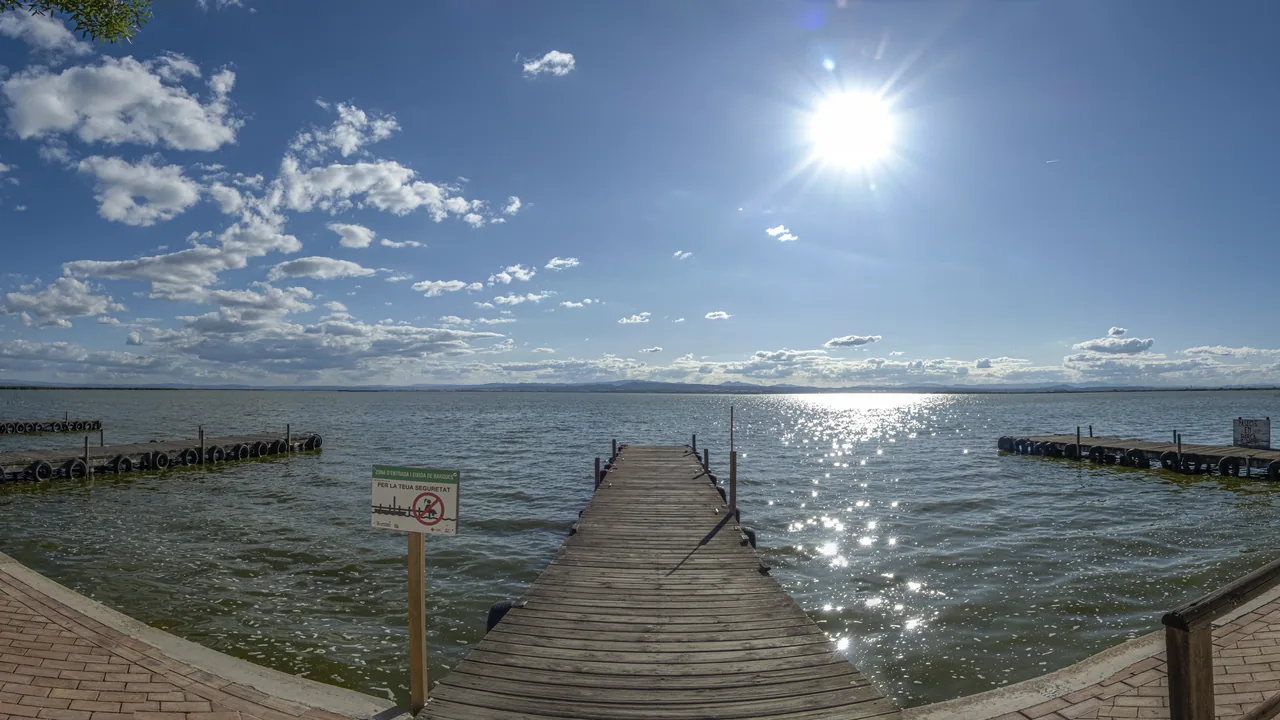 Un embarcadero de madera adentrándose en la laguna de la Albufera bajo un sol reflejado en el agua.